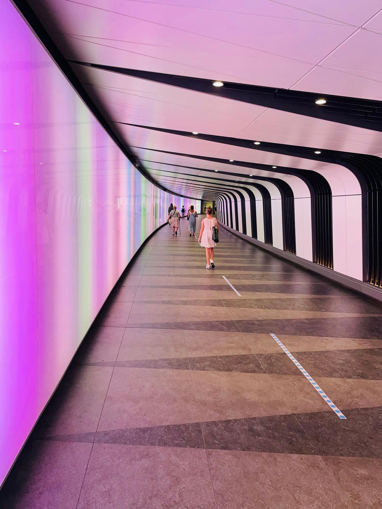 A long curved walkway lit in changing colours, with a few people walking in the distance