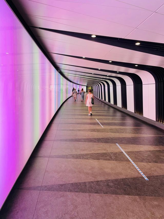 A long curved walkway lit in changing colours, with a few people walking in the distance