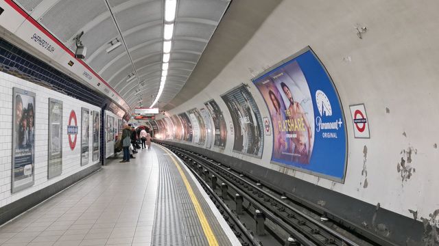 A curved Central line platform at Shepherd's Bush with a platform sign, posters on the tunnel wall and a few passengers waiting