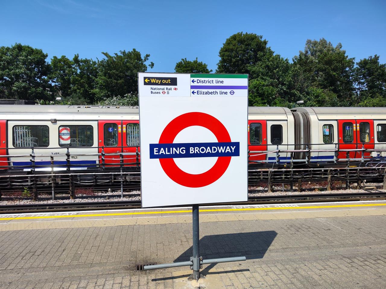 An Ealing Broadway roundel sign on a platform, with direction signs for the District and Elizabeth lines and a District line train behind