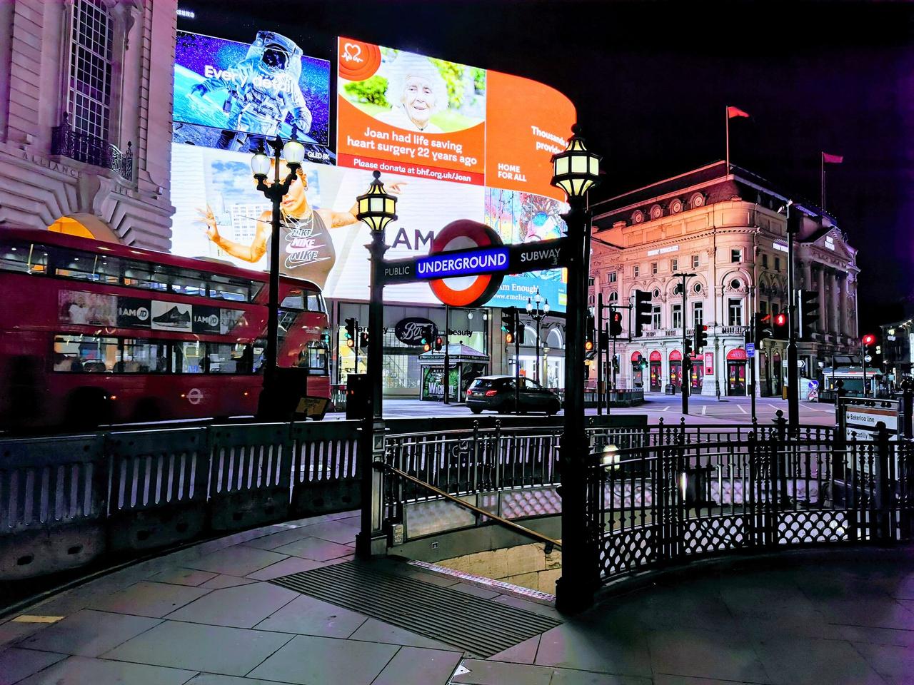 An Underground roundel on a lamp post above the railed stairway entrance at Piccadilly Circus at night, with lit advertising screens and a red bus behind