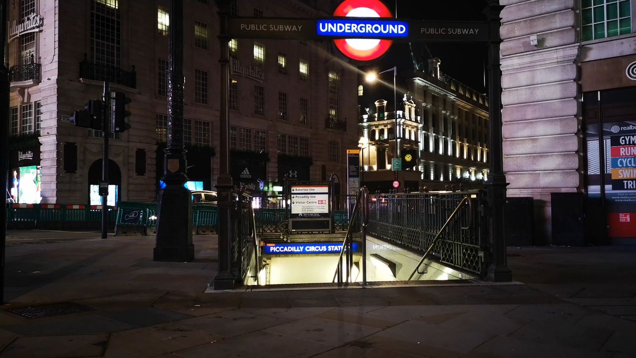 An Underground roundel sign above a lit stairway entrance marked Piccadilly Circus Station, on a quiet street at night